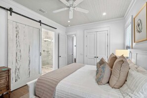 Une chambre à coucher comprenant un lit, un ventilateur de plafond et une porte coulissante donnant accès à une salle de bain.