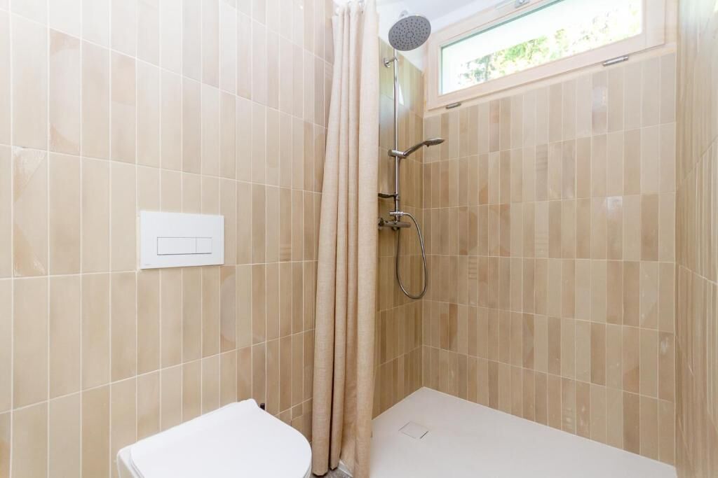 Ein Badezimmer mit beigen Fliesen, einer Dusche inklusive Duschbrause und einem Fenster.