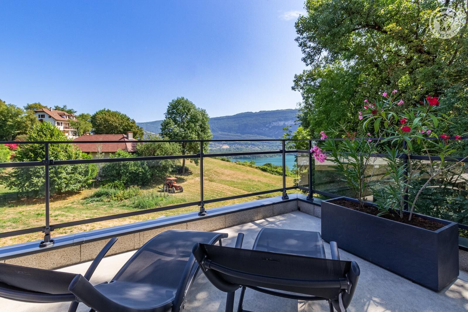 A balcony with a view of a lake and mountains, a black chair, and a potted plant with pink flowers.