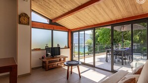 Une pièce avec un plafond en bois, un téléviseur posé sur un meuble en bois, un coin repas avec une table et des chaises, et de grandes fenêtres donnant sur un lac et la verdure environnante.