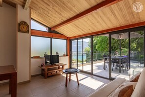 Ein Zimmer mit Holzboden, einem Fernseher auf einem Holztisch, einer Essecke mit Tisch und Stühlen sowie großen Fenstern mit Blick auf einen See und Grünflächen.