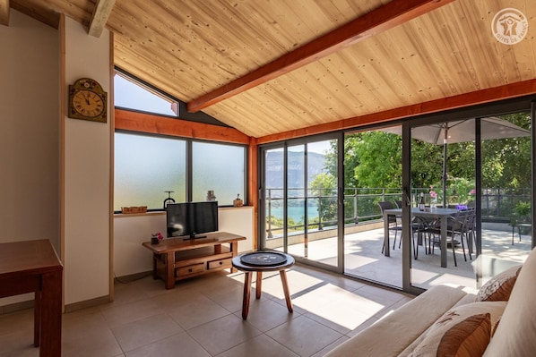A room with a wooden ceiling, a television on a wooden stand, a dining area with a table and chairs, and large windows with a view of a lake and greenery. - The Gîte De La Marquise (CONJUX)