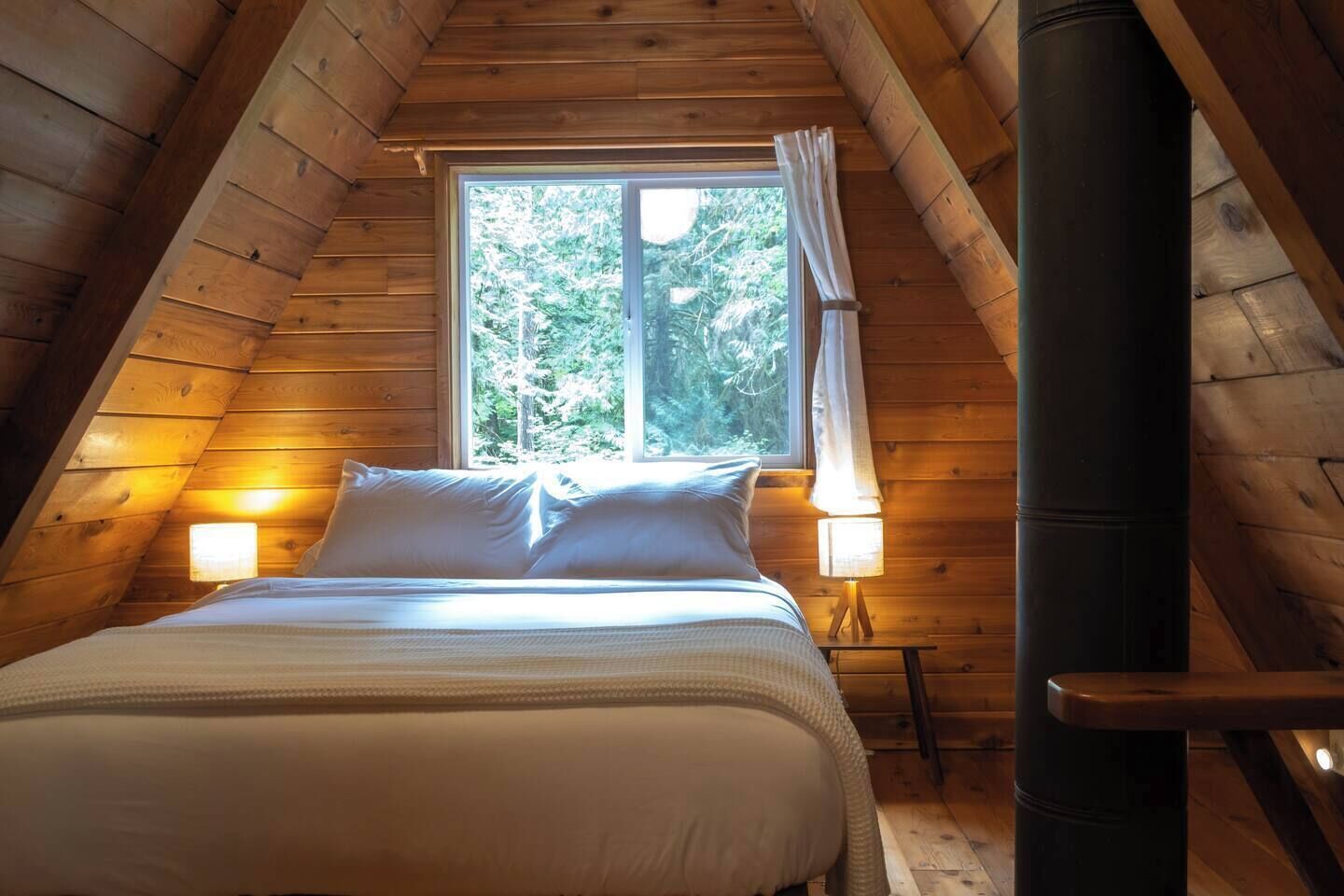 Un dormitorio con techo inclinado, una cama con ropa de cama blanca, dos lámparas de noche y un ventanal con cortinas.