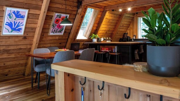 A cozy cabin interior with a dining area, kitchen, and a large window. - Forest A-Frame & Sauna : Dog Friendly + Pool (Deming)