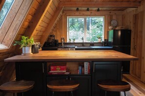 A kitchen with a wooden countertop, a sink, a stove, and a window. There are three stools and a shelf with books. - Forest A-Frame & Sauna : Dog Friendly + Pool (Deming)