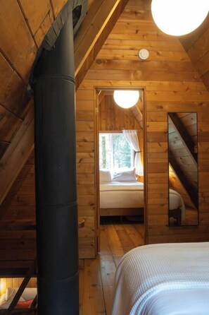 A wooden cabin interior with a bed, a visible chimney, and a window with curtains. - Forest A-Frame & Sauna : Dog Friendly + Pool (Deming)