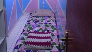 A small, single-bed room with a geometric patterned bedspread, a single wall-mounted light, and a door on the right.