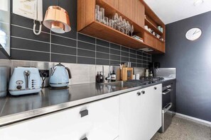 A modern kitchen with a black tiled backsplash, white cabinets, a stainless steel countertop, a kettle, a toaster, and a wooden shelf with glassware. - Private balcony garden oasis in the heart of Fitzroy (Fitzroy)