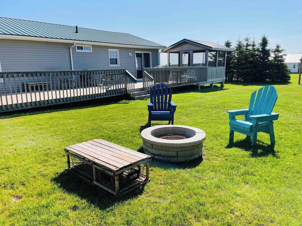A grassy outdoor area with a fire pit, two Adirondack chairs, and a wooden table.