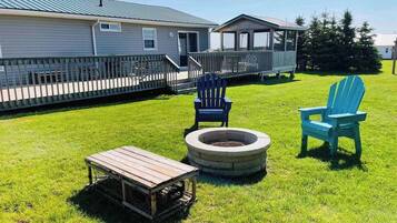 A grassy outdoor area with a fire pit, two Adirondack chairs, and a wooden table.