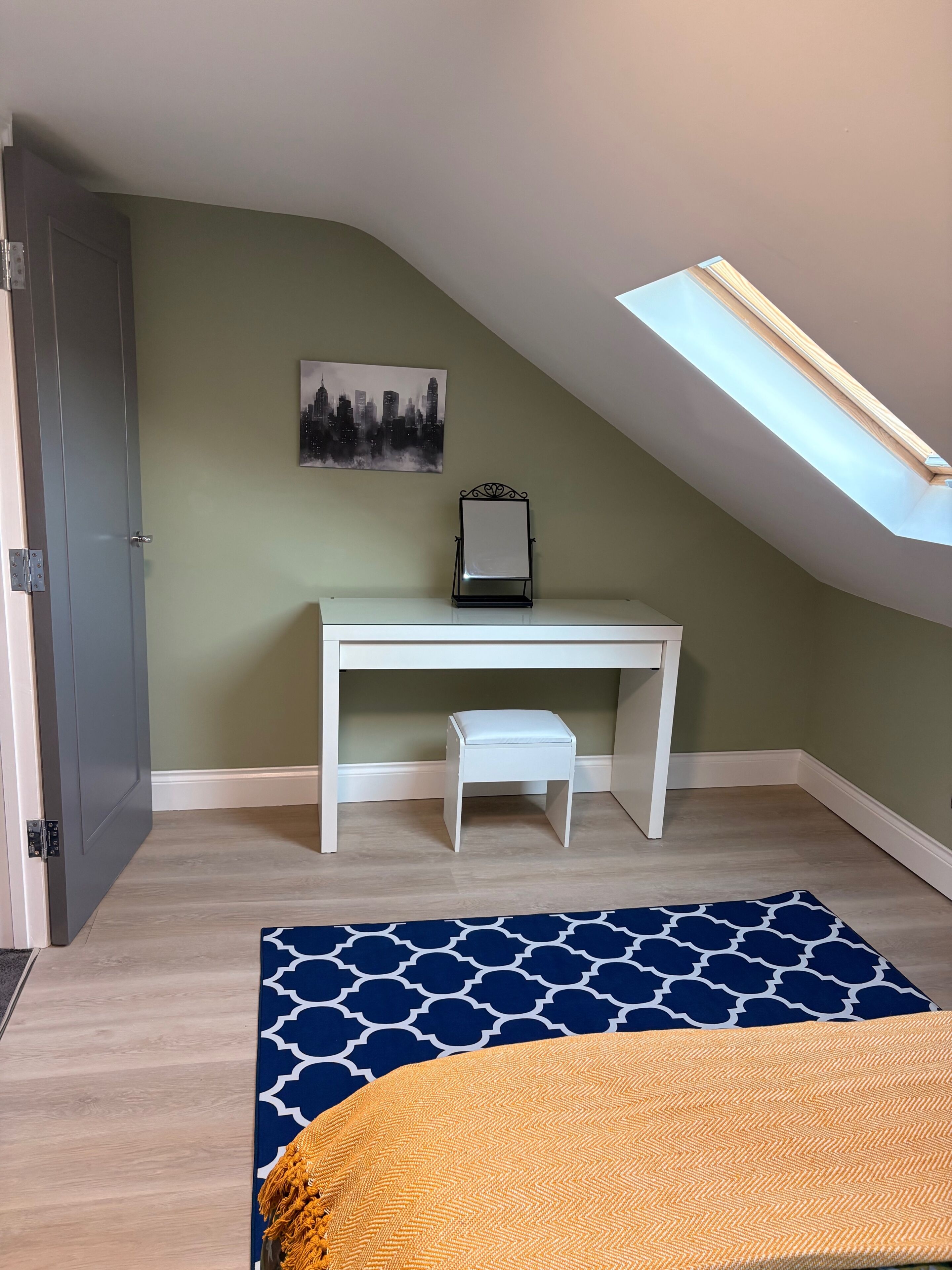 Ein Zimmer mit schrägem Dach, einem Schreibtisch mit Stuhl, einem Bett mit gemustertem Überwurf und einem Stadtbild an der Wand.