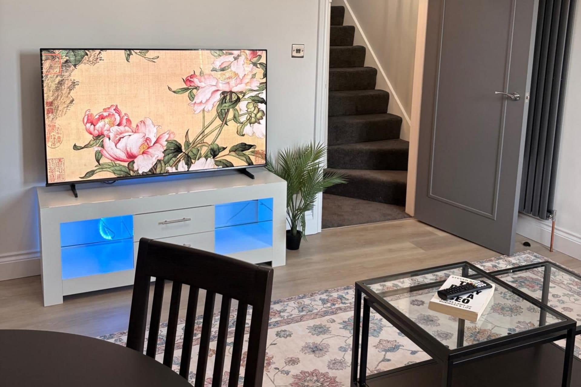 Una sala de estar con una televisión de pantalla plana sobre un mueble blanco, una mesa de centro de vidrio con un libro, una silla de madera oscura y una escalera al fondo.