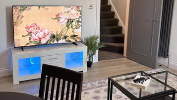 Una sala de estar con una televisión de pantalla plana sobre un mueble blanco, una mesa de centro de vidrio con un libro, una silla de madera oscura y una escalera al fondo.