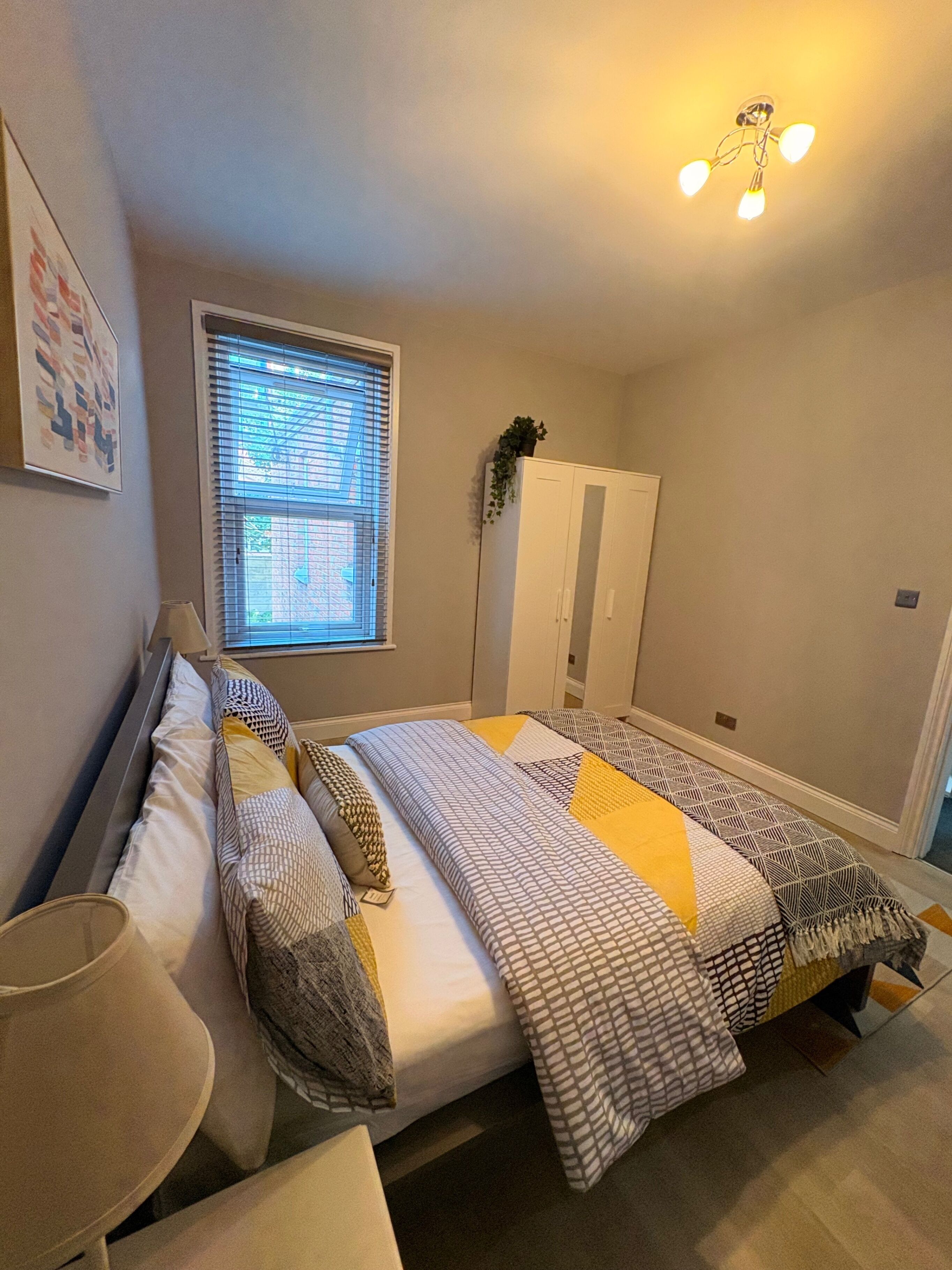 Ein Schlafzimmer mit Bett, Nachttisch, Lampe, Fenster und Kleiderschrank.