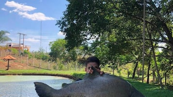 Uma pessoa segurando um peixe grande perto de um corpo d'água, com árvores e uma cerca ao fundo.