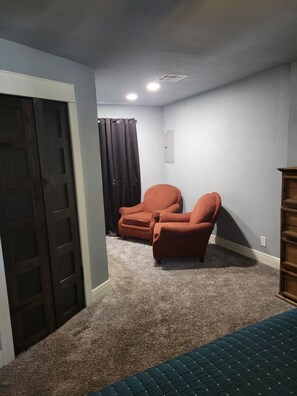 A room with carpeted flooring, a door, two chairs, a ceiling light, and a wall-mounted shelf. - Spacious 4-bedroom house in Muskogee with AC (Muskogee)