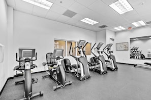Un gimnasio con varias bicicletas estáticas y cintas de correr, paredes blancas y un espejo grande.