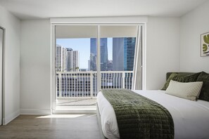 Quarto com cama grande, vista de edifícios altos por uma porta deslizante e varanda com guarda-corpo.