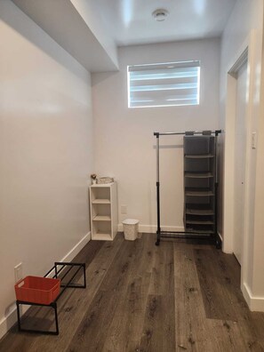 A hallway with a wooden floor, a white shelf unit, a red basket on a black stand, a window with blinds, and a black metal rack. - A Place To Thrive. (Winnipeg)