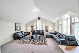 A spacious living room with a high ceiling, large windows, and a mix of sofas and a coffee table.