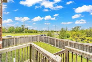 A fenced outdoor area with a grassy lawn, a wooden deck, and a view of a parking lot with cars.