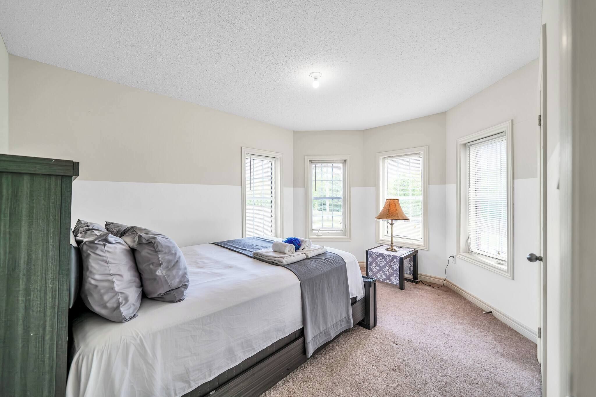 A bedroom with a bed, pillows, a bedside table with a lamp, and windows.