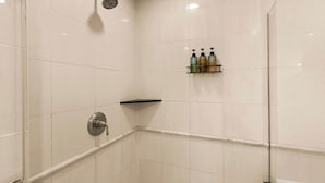 Kylpyhuone, jossa on valkoiset laatat, suihkupää, wc-istuin ja hylly, jossa on hygieniatuotteita.