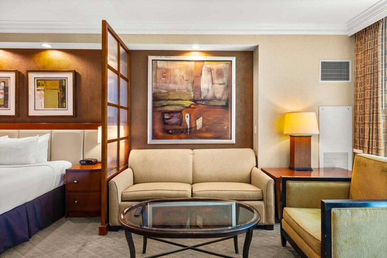 Habitación de hotel con cama, sofá, mesa de centro de vidrio, un cuadro en la pared y una lámpara.