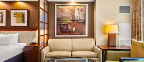 Habitación de hotel con cama, sofá, mesa de centro de vidrio, un cuadro en la pared y una lámpara.