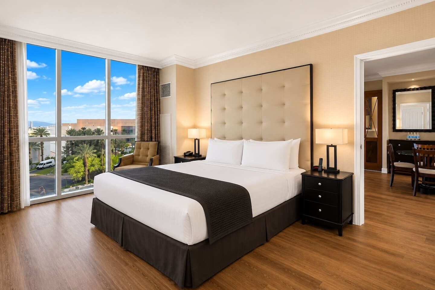A hotel room with a large bed, two bedside tables, a desk with chairs, and a view of palm trees and a parking lot.