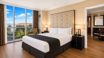 A hotel room with a large bed, two bedside tables, a desk with chairs, and a view of palm trees and a parking lot.