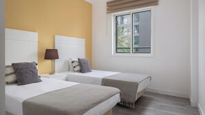 A hotel room with two beds, a window with blinds, a lamp, and a view of a building outside. - Ajuda VI – Comfortable Apartment in Funchal (Funchal)