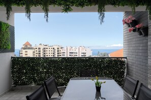 A balcony with a view of a cityscape and the sea, featuring a table and chairs, a potted plant, and hanging flower pots. - Ajuda VI – Comfortable Apartment in Funchal (Funchal)