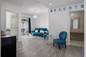 Ein modernes Wohnzimmer mit einem blauen Sofa, einem kleinen runden Tisch und einem blauen Sessel. An der Wand ist ein Schild mit der Aufschrift 'WELCOME' sichtbar.