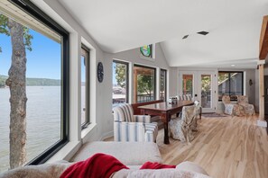 A spacious living area with a large window overlooking a body of water, a dining area with a wooden table, and a cozy seating arrangement. - A Stunning east facing, lake front home, in a cove, with Main Channel Views! (Sunrise Beach)
