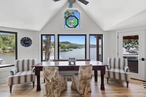 A dining area with a wooden table and striped chairs, large windows with a view of a lake and boats, a clock on the wall, and a stained glass window above. - A Stunning east facing, lake front home, in a cove, with Main Channel Views! (Sunrise Beach)