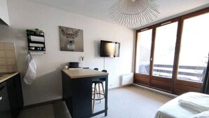 A hotel room with a bed, a desk with a chair, a television, a wall-mounted shelf with towels, and a large window. - Studio cosy avec coin cabine – Le Sauze (Enchastrayes)
