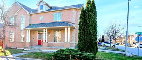 A two-story brick house with a red front door, a covered porch, and a tall evergreen tree in the front yard.
