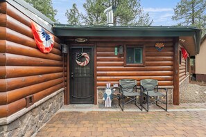 A log cabin with a black door, a welcome sign, a number sign, and two chairs outside. - The Blue Spruce Log Cabin | Cozy 3BR | Munds Park (Munds Park)