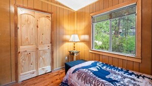 Un dormitorio con paneles de madera, una cama con colcha estampada, un ventanal con persianas y una mesita de noche con lámpara.