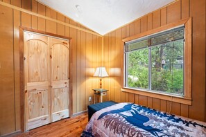 Ein Schlafzimmer mit Holzwänden, einem Bett mit gemustertem Überwurf, einem Fenster mit Jalousien und einem Nachttisch mit Lampe.
