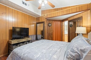 Een slaapkamer met houten lambrisering, een flatscreen televisie, een bed met bloemenprint beddengoed, een nachtkastje met een lamp, en een deur die naar een andere kamer leidt.