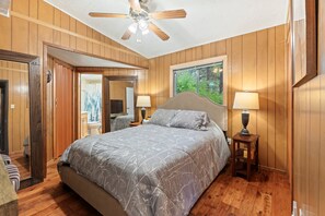 A bedroom with a bed, a nightstand, a window with a view of greenery, and a ceiling fan. - The Blue Spruce Log Cabin | Cozy 3BR | Munds Park (Munds Park)