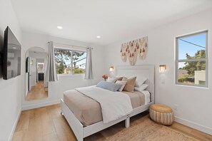 A modern bedroom with a bed, bedside lamps, a TV, and a large window. - Spacious Coastal Getaway. Walk to Windansea beach, coffee shops, restaurants. (La Jolla)