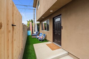 A narrow backyard with artificial grass, a wooden fence, a door, and a small seating area with a chair and a table. - Spacious Coastal Getaway. Walk to Windansea beach, coffee shops, restaurants. (La Jolla)