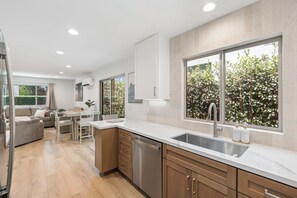 A modern kitchen with a large window, a built-in dishwasher, and a dining area with a table and chairs. - Spacious Coastal Getaway. Walk to Windansea beach, coffee shops, restaurants. (La Jolla)