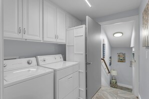 A laundry area with two front-loading washers and dryers, a door leading to another room, and a hallway with a lamp and wall art.