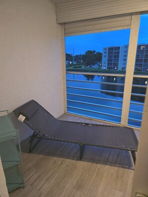 A room with a single reclining chair, a window with a view of a building across a body of water, and a metal shelf unit. - Spacious Dania Beach Condo Minutes from Beach, Airport & Cruise Port (Dania Beach)