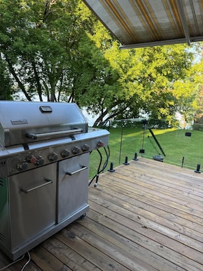 Un barbecue en acier inoxydable doté de plusieurs brûleurs et d’un four intégré, installé sur une terrasse en bois et surplombé d’une zone abritée.