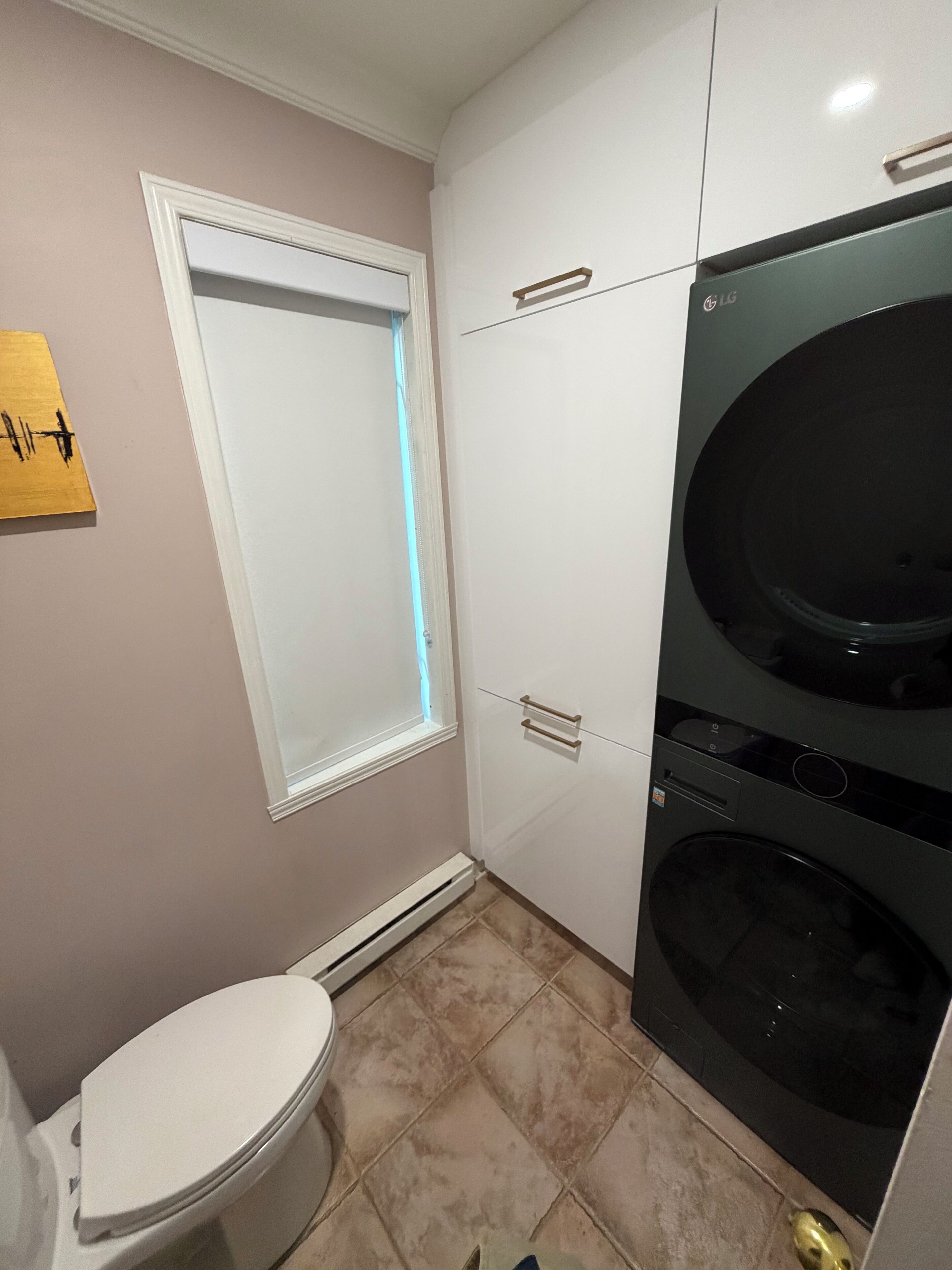 A compact bathroom with a toilet, a window with blinds, and a washing machine.